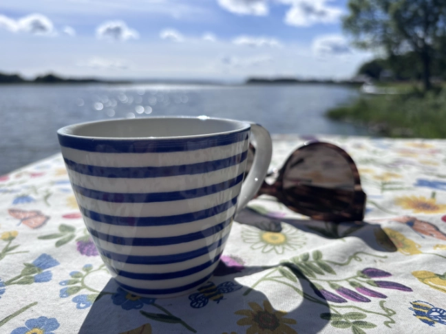 Randig kaffekopp och solglasögon på en blommig duk framför en glittrande sommarsjö.